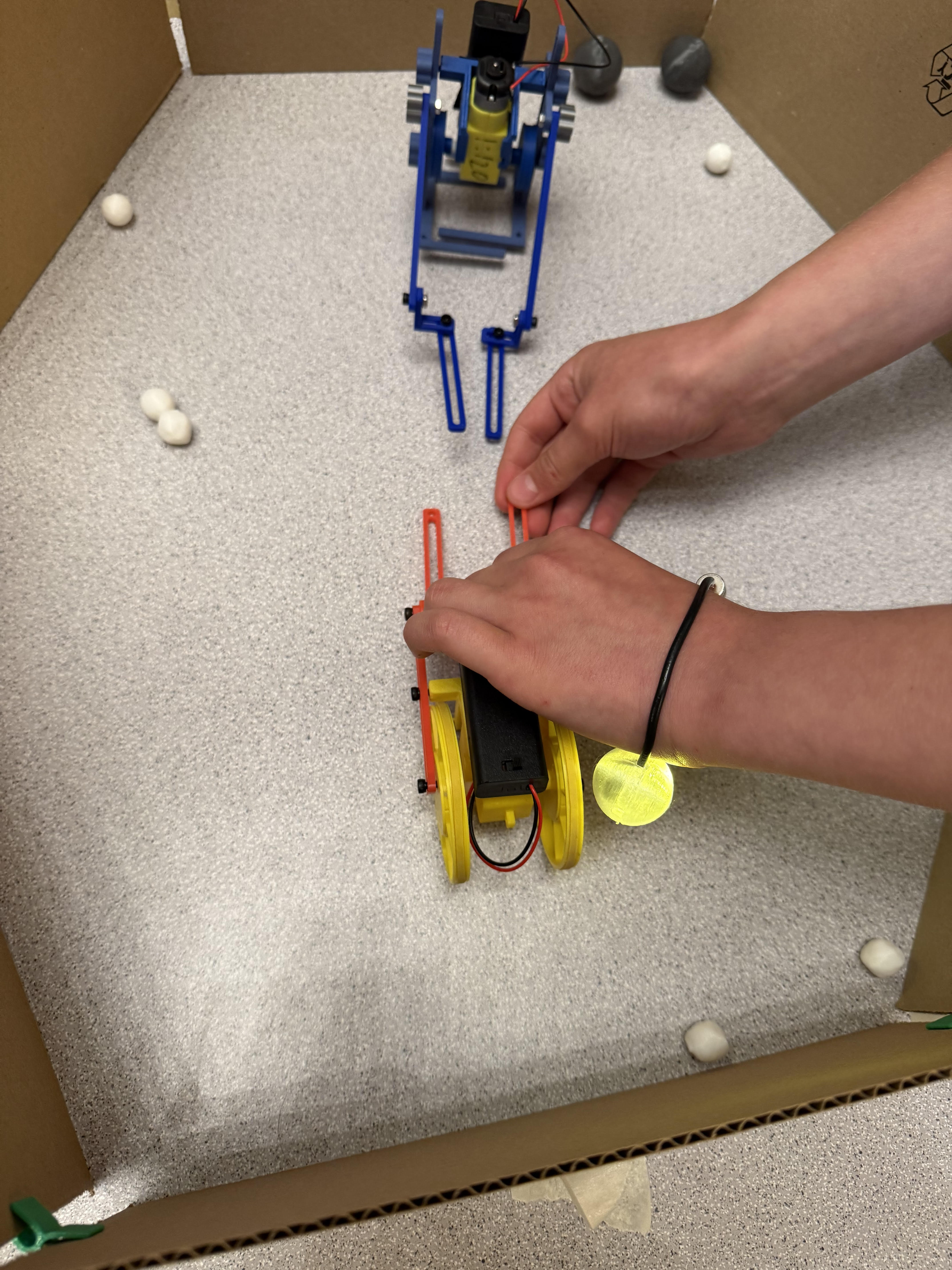 Two robots facing off inside the cardboard arena, a hand steering the yellow wheeler