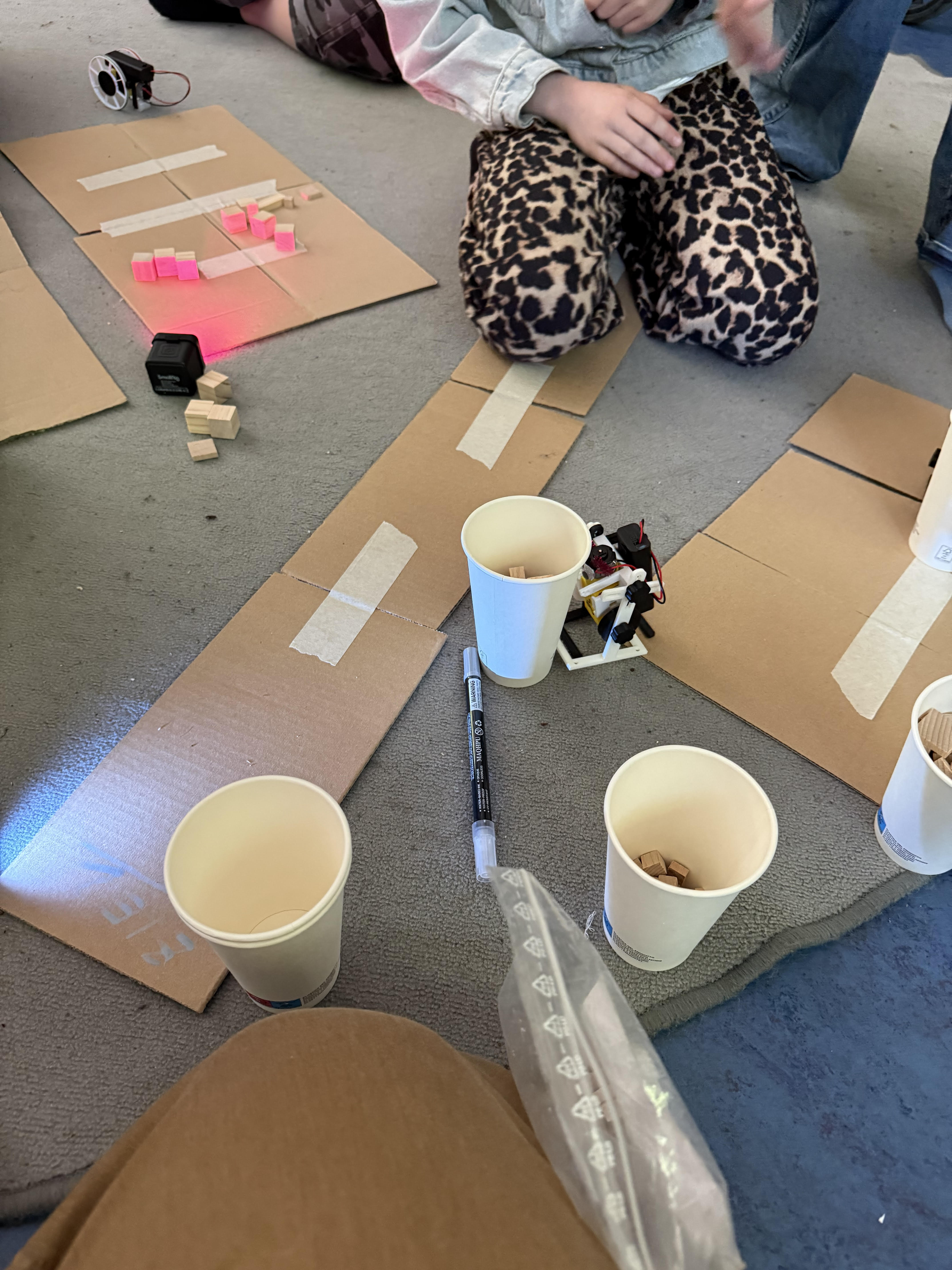 A robot navigating a cardboard obstacle course on the floor with paper cups and wooden blocks
